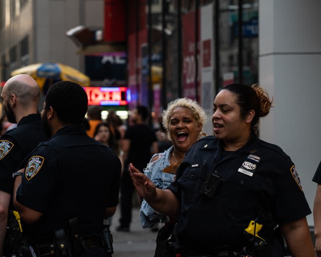 Police officers are engaged in a conversation on a busy street, with one officer smiling broadly. The background features city life with pedestrians and illuminated signs