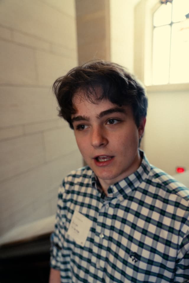 A young person with short hair, wearing a checkered shirt and a name tag, stands indoors with a window in the background, looking slightly to the side