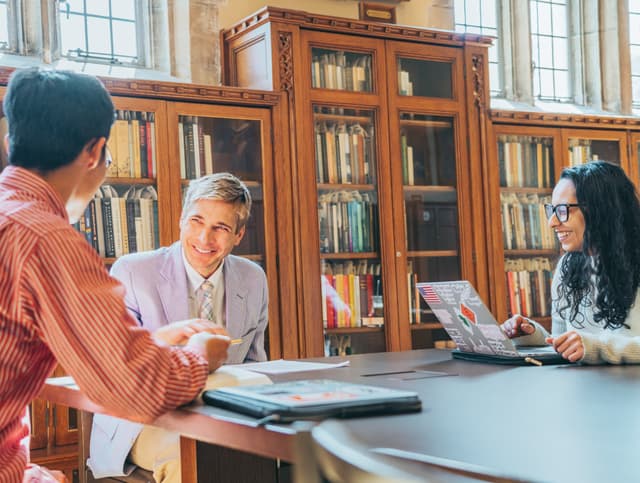 Three individuals are engaged in a discussion at a round table in a library, surrounded by bookshelves filled with books They appear to be in a collaborative and cheerful conversation, with one person holding a document or book and another using a laptop The setting suggests an academic or research environment