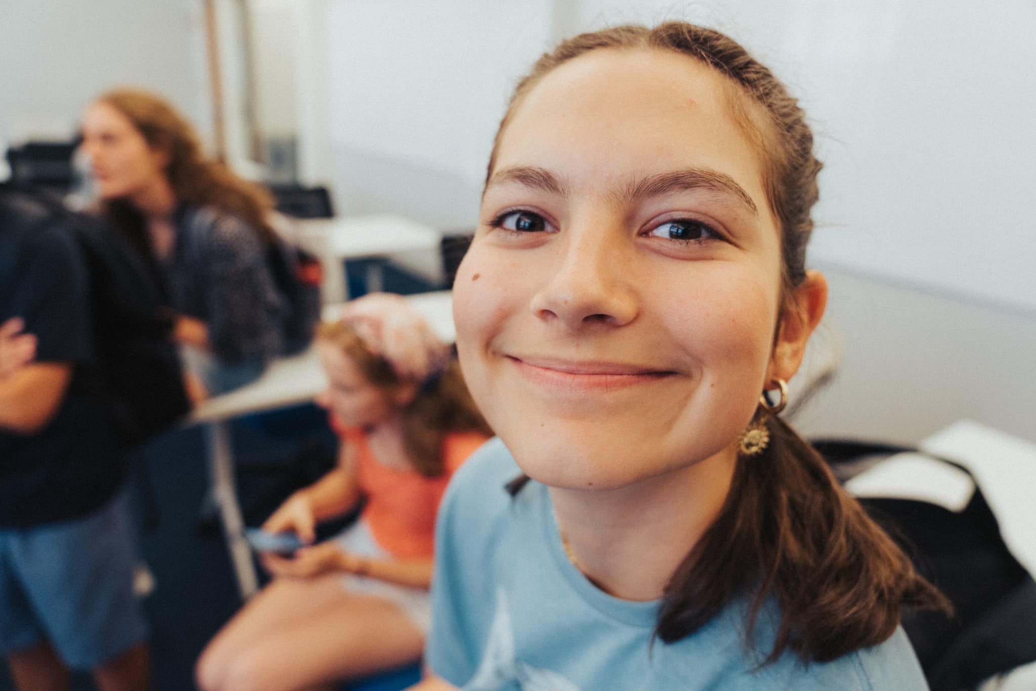 A smiling young woman in a blue shirt is in focus in the foreground, with other individuals and classroom elements slightly blurred in the background