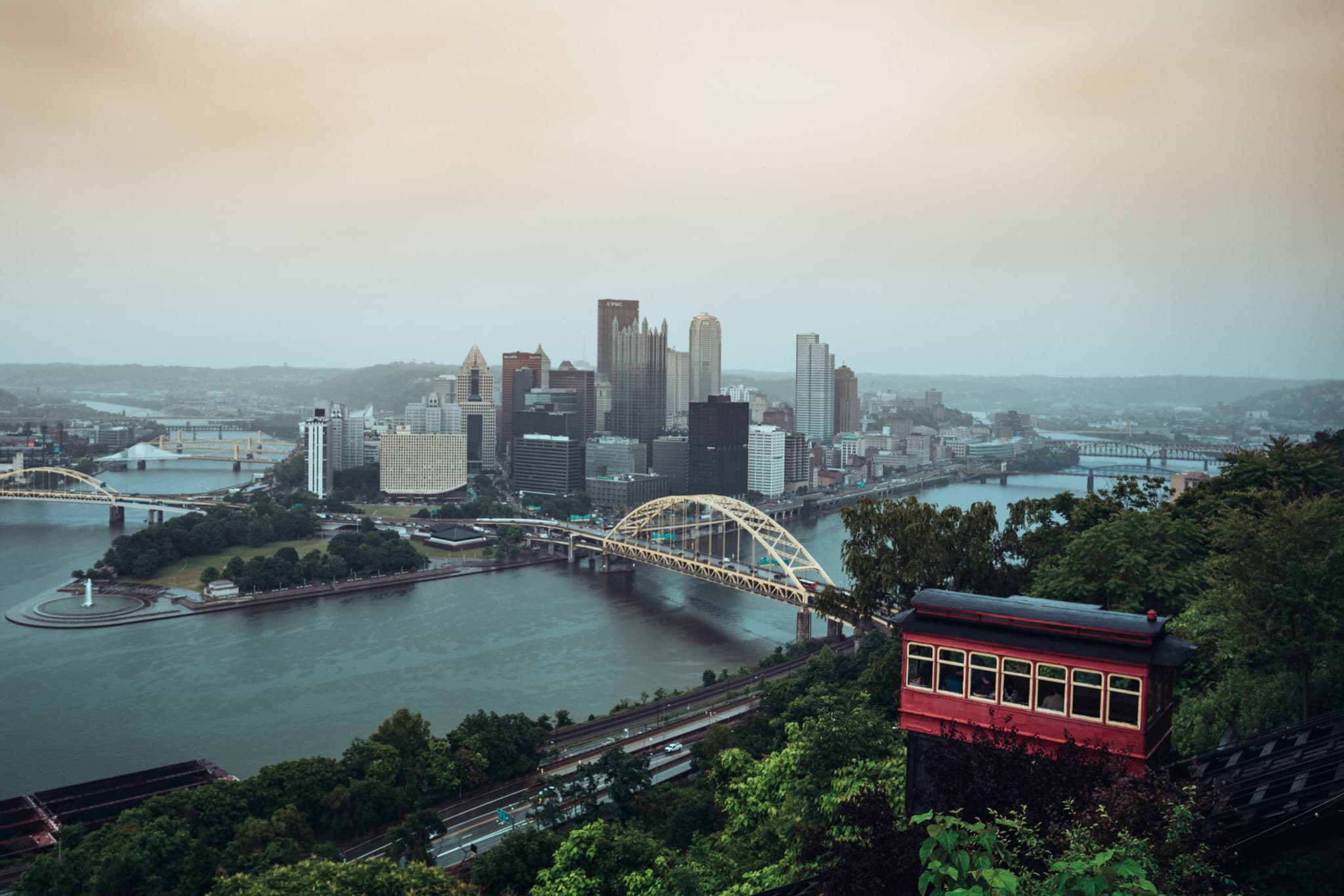 A panoramic view of a city skyline at dusk with a river, a distinctive yellow bridge, and an incline railway car in the foreground