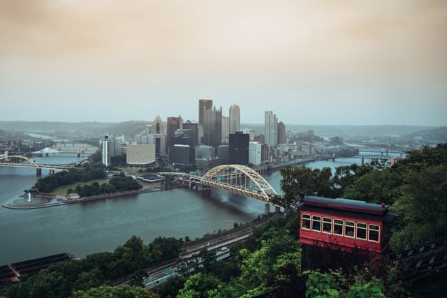 A panoramic view of a city skyline at dusk with a river, a distinctive yellow bridge, and an incline railway car in the foreground