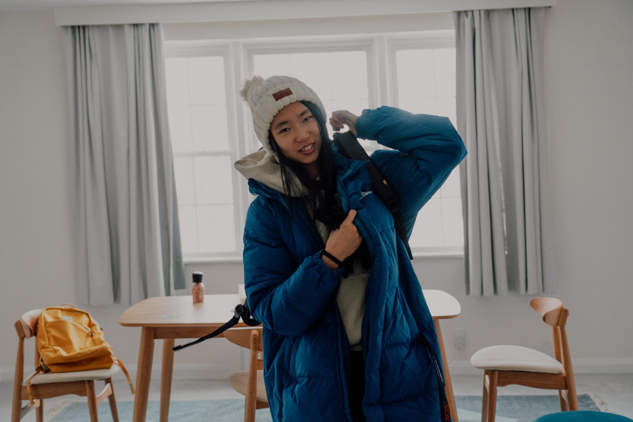 A person in winter attire, including a beanie and a blue coat, is smiling and adjusting their coat indoors, with a wooden table and chairs, and a yellow backpack in the background