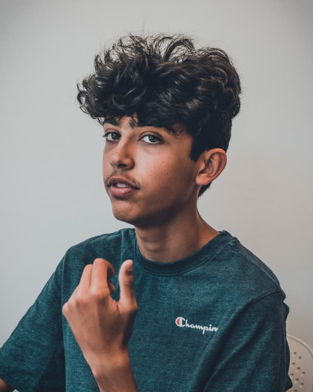 A young person with curly hair wearing a Champion t-shirt is making a hand gesture, possibly beckoning or making a point, while looking slightly to the side