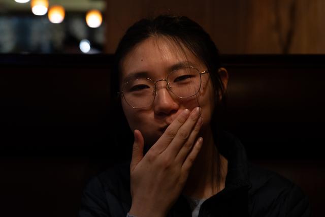 A smiling woman with glasses covering her mouth with her hand, in a dimly lit environment with soft background lights