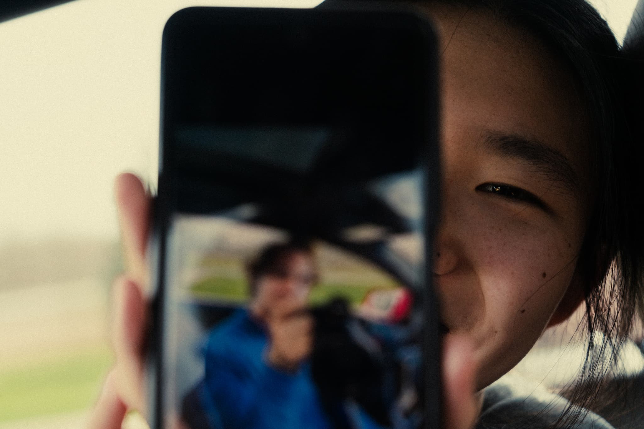 A smiling person is partially visible behind a smartphone, which displays the reflection of another person wearing a blue jacket