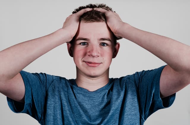 A young man in a blue t-shirt is holding his head with both hands, looking slightly stressed or perplexed, against a neutral background