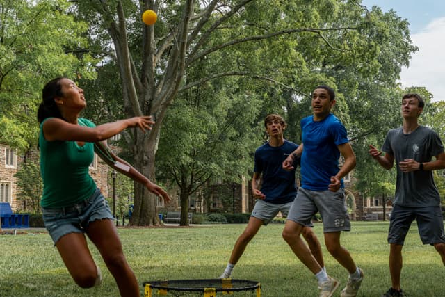 Four people are playing a game of spikeball outdoors on a grassy field with trees in the background. One player is hitting the ball towards the small, round net in the center while the others watch in anticipation