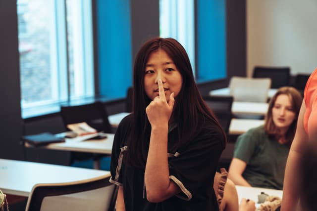 A young woman in a classroom setting appears to be listening intently, with her hand near her mouth, possibly indicating thoughtfulness or concentration. Other students are visible in the background, suggesting an educational environment