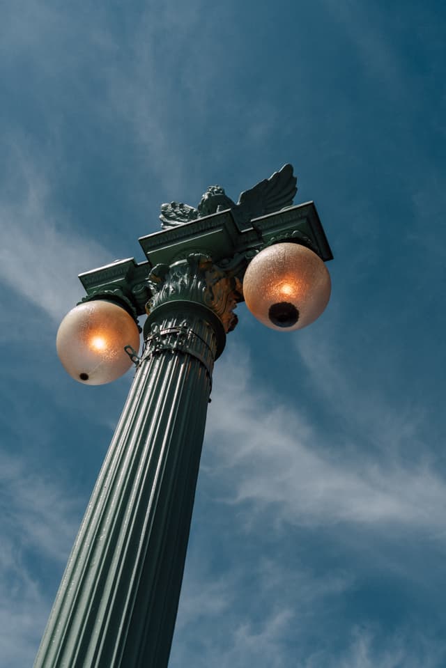 A tall, ornate street lamp with two glowing globes against a blue sky with wispy clouds