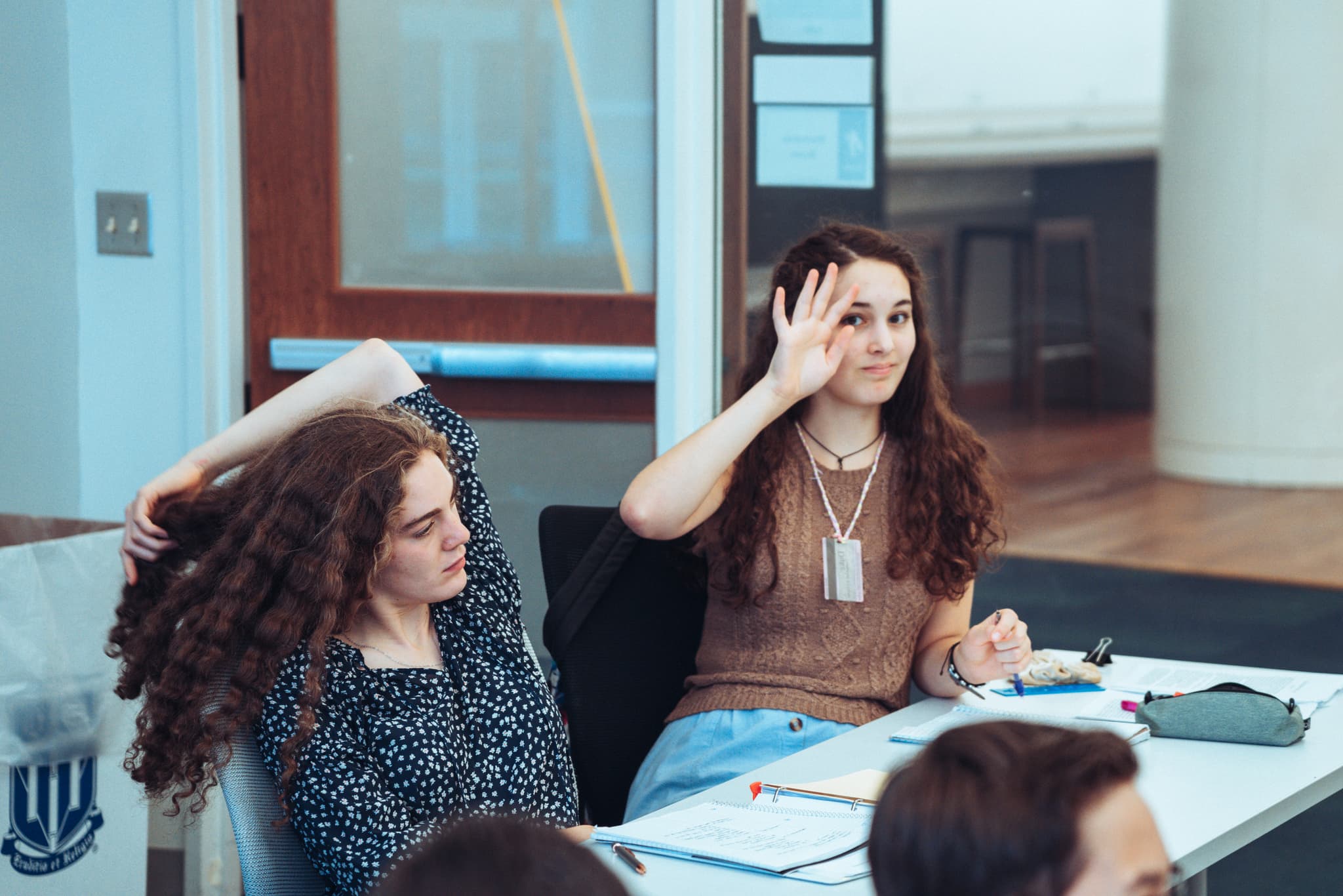 Two women seated at a table appear to be in a classroom or meeting setting, with one raising her hand as if to ask a question or make a comment. They are surrounded by papers and notebooks, suggesting an educational or professional environment