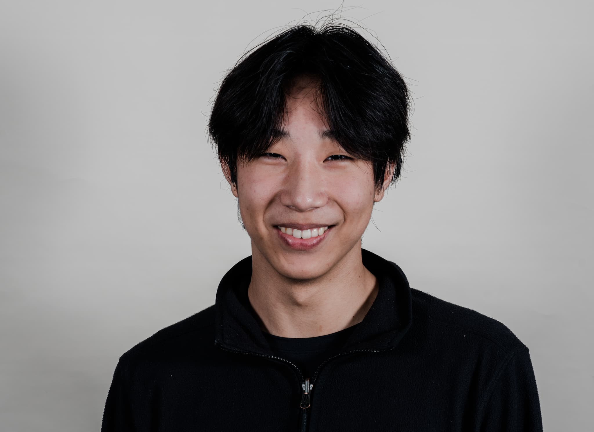 A smiling man with black hair wearing a black zip-up top against a grey background